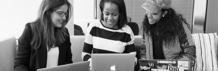 Three recially diverse women sitting on a sofa each with a
laptop on their lap and smiling.